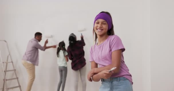 Renovación de apartamento chica sonriente en una blusa púrpura y diadema mira en la cámara sostiene un pincel en la mano ríe en el fondo una familia está pintando paredes junto a una escalera — Vídeos de Stock