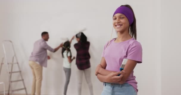 Remont mieszkania uśmiechnięta dziewczyna w fioletowej bluzce i opasce patrzy na aparat z dumą trzymając pędzel w tle rodzina maluje ściany obok drabiny — Wideo stockowe