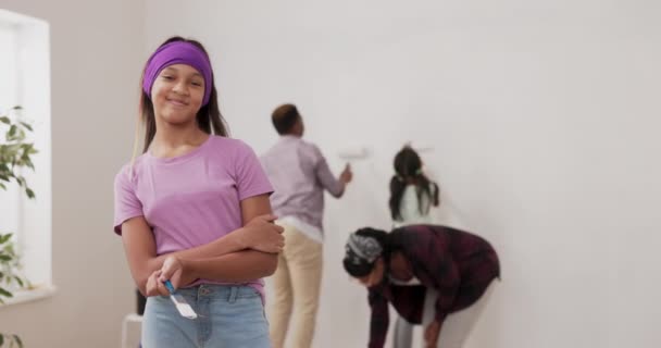 Rénovation de l'appartement une jeune fille souriante dans un chemisier violet et bandeau regarde la caméra fièrement tenant le pinceau à la main en arrière-plan la famille peint les murs à côté de l'échelle — Video