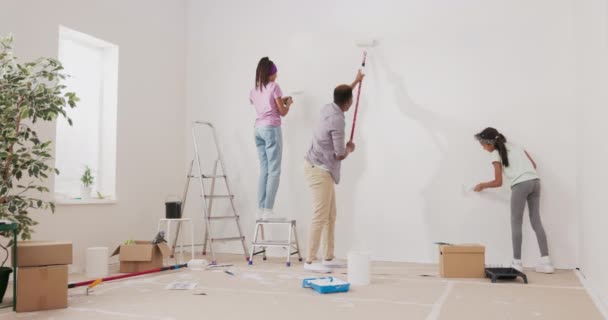 En ung far och döttrar renoverar en lägenhet i avsaknad av hans fru överraskning de håller rullar med vit färg och exakt måla väggarna en flicka klättrar en stege — Stockvideo