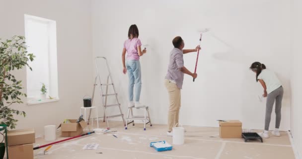Un joven padre e hijas renuevan un apartamento en ausencia de su esposa sorpresa sostienen rodillos con pintura blanca y pintan con precisión las paredes una niña sube una escalera — Vídeos de Stock