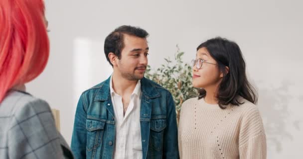 Happy couple stand in empty room while inspecting apartment for rent with real estate agent woman and man look at place indicated by pink-haired girl touring rooms showing out equipment — Stock videók