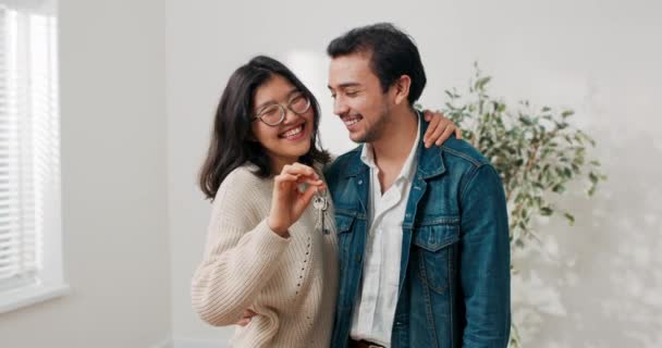 Jong stel in de liefde huren van hun eerste appartement samen vrolijke vrouw omarmt vriendje in de hand houden sleutels van flat ze staan in het midden van een onlangs gerenoveerde lege kamer — Stockvideo