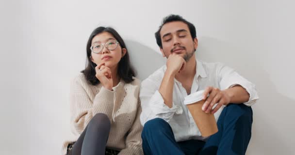 Una pareja enamorada está sentada junto a la pared en un apartamento nuevo, están mirando alrededor de la habitación pensando profundamente en la elección de la decoración, amueblando la casa, bebiendo café de tazas para llevar — Vídeo de stock