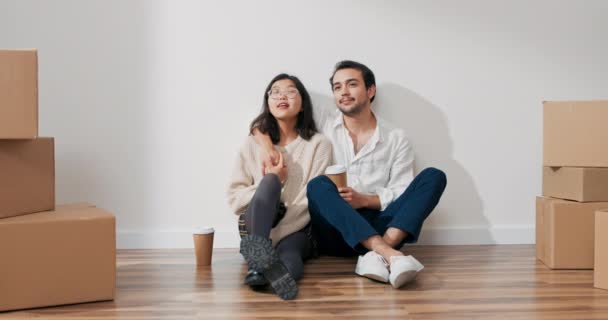 Una pareja está sentada junto a la pared en un apartamento nuevo, un hombre galante vestido con una camisa blanca está charlando con una hermosa mujer sonriente, están tomando café, hablando del aparato, decoración plana — Vídeo de stock