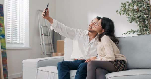 Una pareja casada, recién trasladada a un apartamento comprado, están sentados en el sofá, abrazándose, el hombre se está tomando una selfie con su amada, los amantes son felices, sonriendo — Vídeo de stock