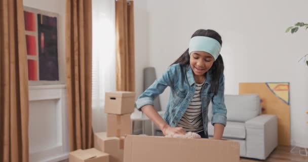 Lány segít a szüleinek kicsomagolni dobozok költözés után talál kartondoboz a játékait húzza ki Teddy maci boldog ölelés szorosan boldog az új otthon — Stock videók