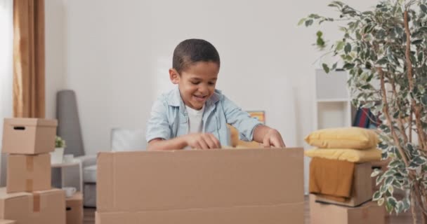 Un niño lindo desempaca su caja de juguetes después de mudarse a una nueva casa saca un coche disfruta juega con él — Vídeo de stock