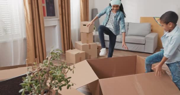 Los hermanos juegan mientras se mueven en cajas de cartón vacías renovación de apartamento nuevo alquiler de una casa que se esconden entre las cosas llenas de los padres alegría niña de la infancia se encarga de niño más joven — Vídeo de stock