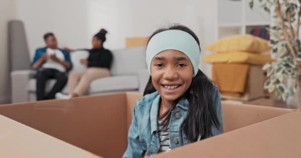 Saç bandı takan sevimli küçük bir kız, taşınma sırasında eşyaları paketlemek için kullanılan karton bir kutunun içinde oturuyor. — Stok video