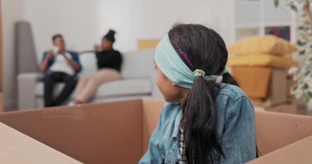 Uma garotinha fofa com uma faixa de cabelo se senta em uma caixa de papelão usada para embalar coisas durante um movimento, irmã se esconde de seu irmão enquanto eles jogam — Vídeo de Stock