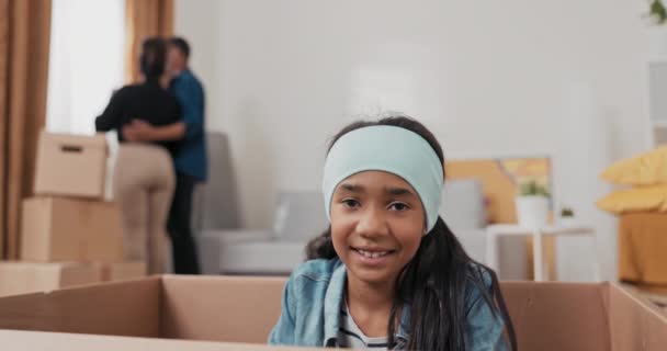 Ein süßes kleines Mädchen mit Haarband sitzt in einem Karton, der zum Packen von Sachen während eines Umzugs benutzt wird. Schwester versteckt sich vor ihrem Bruder, während sie im Hintergrund der Eltern spielen — Stockvideo