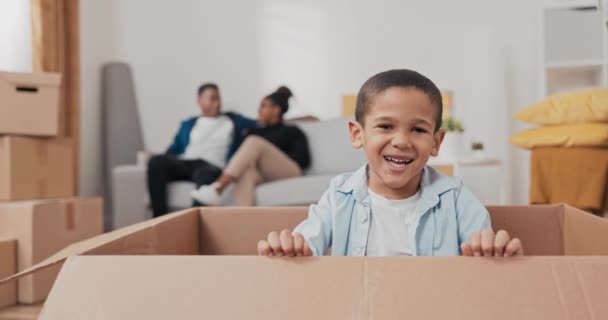 Um menino pequeno bonito sorrindo senta-se em uma caixa de papelão usada para embalar coisas durante um movimento a criança olha para seus pais e depois ri para a câmera — Vídeo de Stock