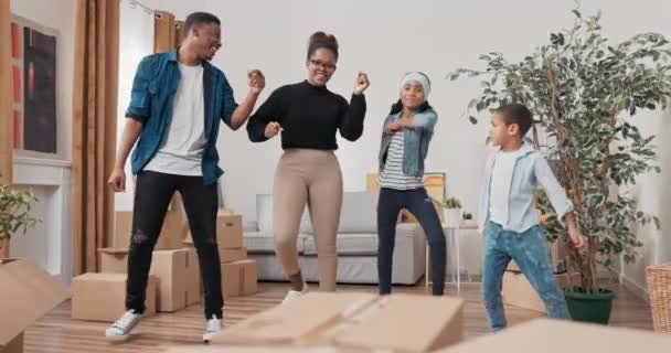 Счастливая семья танцует в середине гостиной молодые родители и их дети празднуют переезд в новую квартиру радоваться вместе практиковать хореографию — стоковое видео