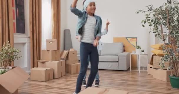 Веселі танець братів і сестер посеред вітальні між картонними коробками в новій квартирі вони переїхали з батьками сестра вчить молодшого брата хореографії — стокове відео