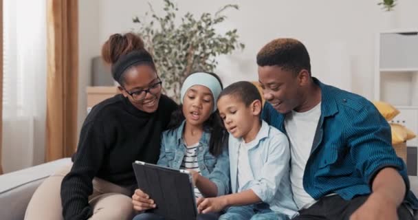 The family spends time together on the couch the parents with their two children browse the furniture for the new apartment on website on tablet the siblings choose decorations for room — Stock Video