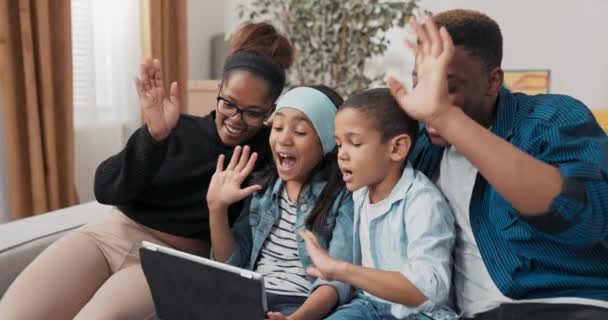 The family is spending time together on the couch the parents and their two children are video chatting with the grandparents on the tablet waving at the camera to say hello talking — Stock Video