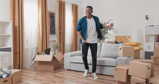 Un homme danse au milieu du salon dans un nouvel appartement gars est heureux de déplacer des boîtes en carton avec des choses emballées se tenir autour de lui joie sourire un nouveau chapitre de la vie — Video