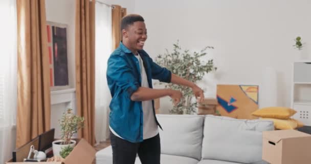 Чоловік, сповнений енергії та оптимізму, танцює в середині вітальні в новій квартирі хлопець із задоволенням рухається навколо нього є картонні коробки з упакованими речами радість посміхається нова глава життя — стокове відео