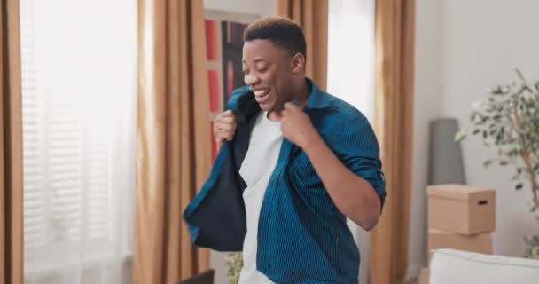 Homem bonito feliz cheio de alegria salta danças no novo apartamento segurando sua camisa caixas de papelão em movimento espalhadas na sala de estar — Vídeo de Stock