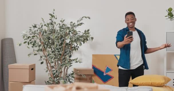 L'homme passe du temps dans l'appartement loué acheté parle au téléphone sur vidéo avec un ami montre ses boîtes en carton décor à la maison rires blagues gestes visite virtuelle — Video