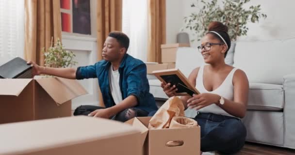 Jóvenes amantes se mudan a un nuevo apartamento se sientan en el suelo y empacan sus cosas en cajas la mujer encuentra un marco con una foto de recuerdo lo miran y recuerdan los viejos tiempos con una sonrisa — Vídeo de stock