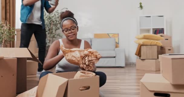 Getrouwd echtpaar verhuist naar nieuw appartement vrouw zit op de vloer uitpakken kartonnen dozen met spullen unrolls items van papier man zet kussens op de bank en zit ontspannen. — Stockvideo