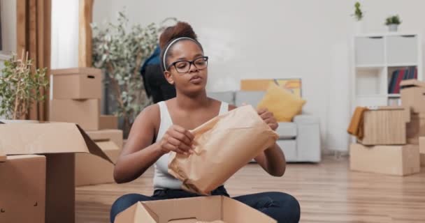 Beautiful woman with glasses sits on floor of purchased rented apartment unpacks cardboard boxes of stuff takes out vase wrapped in paper smiles at camera in background man is cleaning — Stock Video