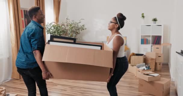 Ένα ερωτευμένο νεαρό ζευγάρι φέρνει βαριά κουτιά με πακεταρισμένα αντικείμενα κορνίζες εικόνων στο αγορασμένο νοικιασμένο σπίτι του κατά τη διάρκεια μιας μετακόμισης ένα χαρτονένιο κουτί στο πάτωμα και κόλλα πέντε. — Αρχείο Βίντεο