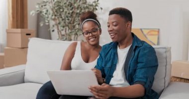 Evli çift, yeni evlerinde kanepede oturuyor. Adam kucağında dizüstü bilgisayarla geziyor. Web sitelerinde mobilya dekorasyonu arıyorlar.