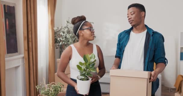 愛のカップルは、彼らが一緒に最初の家を飾る手で植物とポットを保持して装飾を設定し、リビングルームの女性に荷物を詰め段ボール箱を運ぶ新しいアパートに移動します — ストック動画