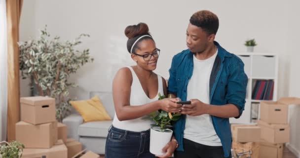 Um casal apaixonado são recém-movido para novo apartamento mulher está de pé no meio da sala de estar segurando planta no homem panela puxa para fora telefone pedindo para tirar selfie eles tiram foto e escolher o melhor — Vídeo de Stock