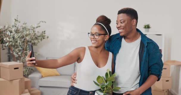 2人の恋は新しいアパートに移される女性は居間の真ん中に立っている彼女のボーイフレンドと一緒に写真を撮る鍋に植物を保持しているソーシャルメディアで — ストック動画