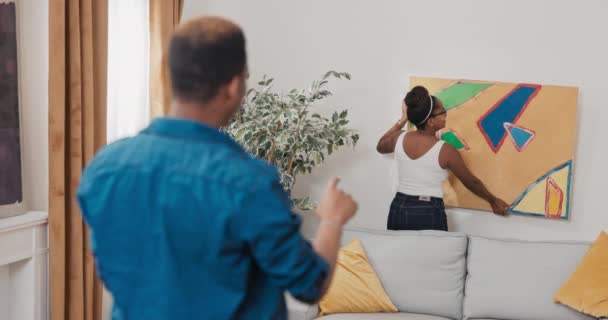 Una mujer cuelga una pintura en la pared en un apartamento nuevo junto con su novio evalúan si se ve bien, el hombre instruye a la chica cómo girar el lienzo de manera que se mueva para que cuelgue uniformemente — Vídeos de Stock