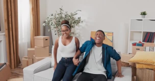 A couple in love tired after moving cardboard boxes into their apartment relax in their newly purchased rented house hold hands and sit down together on the couch to rest — Stock Video