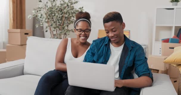 Egy házaspár ül a kanapén az új lakásukban, miután beköltöztek, laptopot tartanak az ölében, és bútorok után kutatnak. — Stock videók