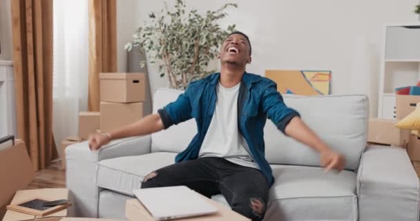 Un uomo siede su un divano da scatole di cartone avanzi da una mossa felice con il suo nuovo appartamento ordinato mobili gioia battendo le mani — Video Stock