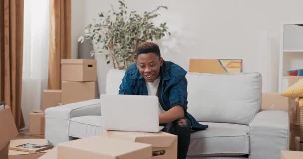 Młody uśmiechnięty chłopiec siedzi na kanapie w nowym mieszkaniu po przeniesieniu sprawdzając e-maile na laptopie dobra wiadomość radość finalizacji zamówienia szczęście ręce w górę zwycięstwo — Wideo stockowe