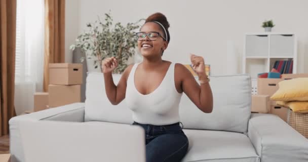 En vacker kvinna med glasögon sitter på en soffa framför en bärbar dator som på pappkartonger i en post-move-in lägenhet goda nyheter seger dans ordning slutförande överföring till konto betalning — Stockvideo