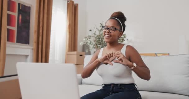En ung kvinna sitter på en soffa i en lägenhet och pratar med sin vän via en bärbar webbkamera de skvallrar skratta flickan tackar uttrycker sina känslor genom att visa ett hjärta från fingrar — Stockvideo