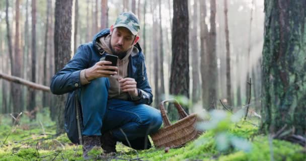 Молодий грибний пікірувальник на коліна в лісі посеред моху і збирає гриби, оглядає знахідку обережно з усіх боків, тримає телефон, в якому перевіряє види, переконується, що це не отруйно — стокове відео