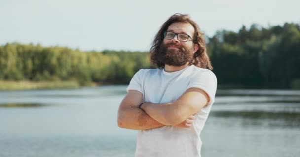 Een portret van een man met lang haar en baard, bril op zijn neus, een jongen steekt armen gekruist, kijkt glimlachend in de camera, zelfverzekerd, op de achtergrond een meer, zomermiddag — Stockvideo