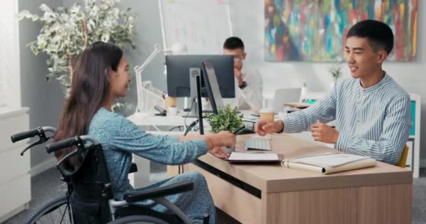 Mulher está sentada em cadeira de rodas na mesa no escritório em todo empregador eles estão assinando contrato empregado homem estende a mão, eles apertam as mãos agradecendo uns aos outros por conversa parabenizando — Vídeo de Stock