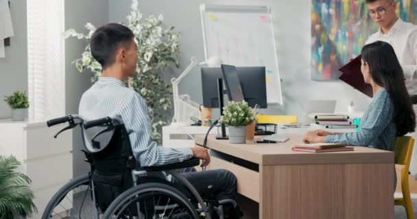 Um homem senta-se em uma cadeira de rodas em uma mesa em um escritório, em frente a ele uma mulher bonita em um vestido, eles assinam um acordo de trabalho cooperativo, agradecer uns aos outros para a entrevista, parabenizar — Vídeo de Stock