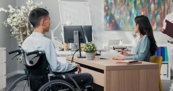 Homens sentam-se em uma cadeira de rodas em uma mesa em um escritório, em frente a ele uma mulher bonita em um vestido, um colega de trabalho traz um contrato eles assinam um documento de colaboração, eles se agradecem pela entrevista — Vídeo de Stock