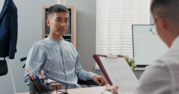 Dos hombres se reúnen en una oficina de la empresa en la mañana para una entrevista de trabajo niño discapacitado llega en silla de ruedas con camisa blanca jefe le entrega un contrato para firmar, felicitaciones apretón de manos por la nueva cooperación — Vídeos de Stock