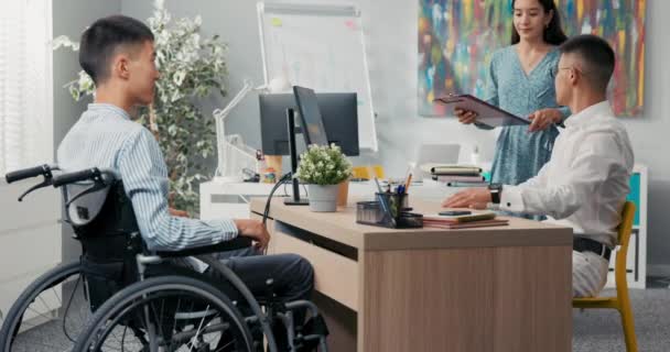 Kobieta sekretarz szef przynosi mu cv kontrakt przyszłego pracownika, niepełnosprawny mężczyzna na wózku inwalidzkim ma rozmowę kwalifikacyjną, kierownik w okularach uśmiecha się dokument znaki daje chłopcu uścisk dłoni gratulacje — Wideo stockowe