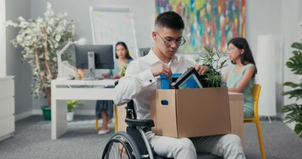 Ung kontorist skadad, olycka, handikappad, i rullstol, håller en låda med paket från sitt skrivbord i knä, pensionering, permitterad, arbetslös, byta plats — Stockvideo