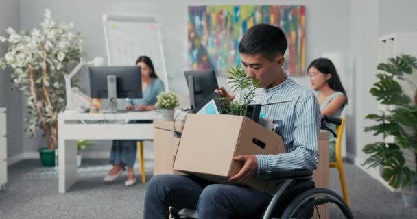 シャツ姿の障害者が社内で就職車椅子を持ったまま職場で働くための必需品を持った箱に座っている同僚の笑顔を見ている — ストック動画