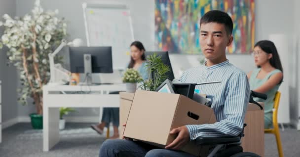 Αναπήρων φιλόδοξος άνθρωπος με πουκάμισο με ασιατική κορεατική ομορφιά κάθεται σε αναπηρική καρέκλα στο γραφείο στην εταιρεία στα γόνατα κρατώντας χαρτονένιο κουτί με συσκευασμένα πράγματα, παίρνει φωτιά από την εργασία σε θέση να εκπληρώσει τα καθήκοντά του — Αρχείο Βίντεο